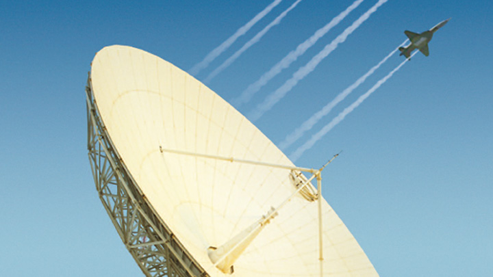 Fotografia di una grande parabola satellitare dietro la quale sfreccia un aereo militare.